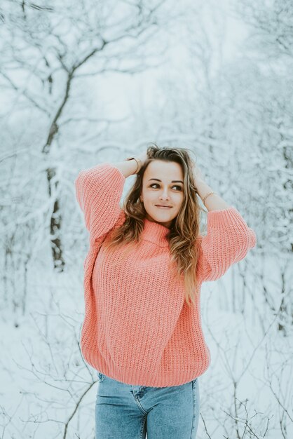 추운 눈 덮인 겨울 숲에서 분홍색 방대한 스웨터와 청바지에 아름 다운 젊은 여자