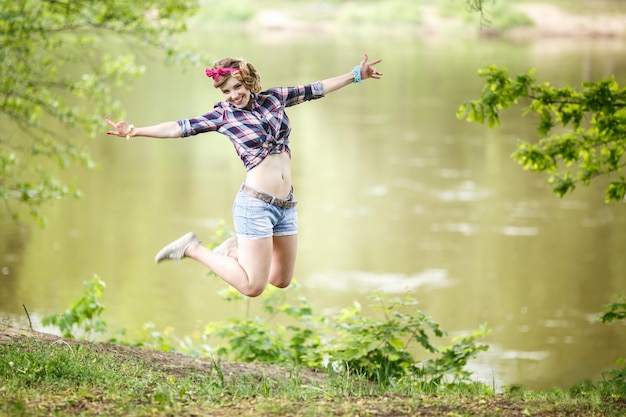 격자 무늬 셔츠와 핀업 스타일의 짧은 데님 반바지를 입은 아름다운 소녀가 숲에서 점프합니다.