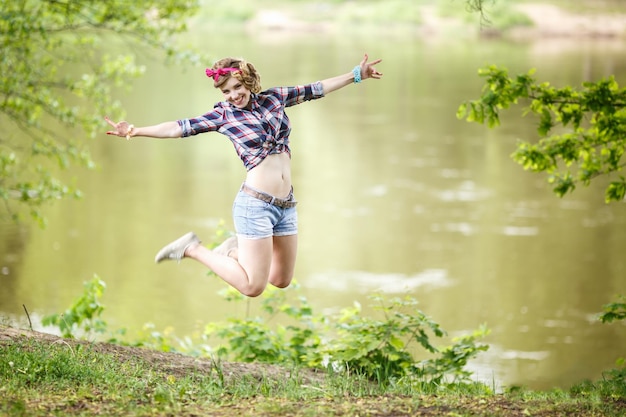 Bella ragazza in una camicia a quadri e pantaloncini di jeans corti in stile pinup salta nella foresta