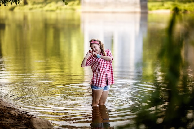 Beautiful young girl in a plaid shirt and short denim shorts in pinup style stays in water of river