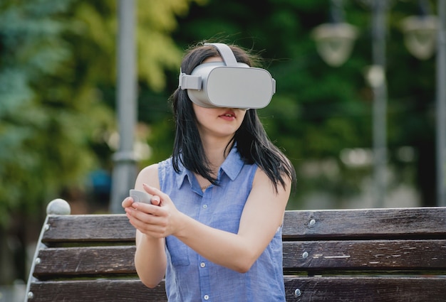 A beautiful young girl plays a game wearing virtual reality glasses on the street.