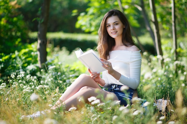 Beautiful young girl reads a book in a summer park outdoors.