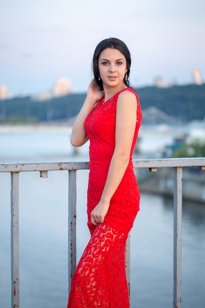 古い柵の近くの橋の上でポーズをとって赤いドレスを着た美しい少女。背景には、焦点がぼけている川、橋、都市の建物の断片があります。
