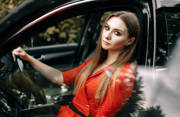 A beautiful young girl in a red overalls sits behind the wheel of a black car on an empty road in the forest