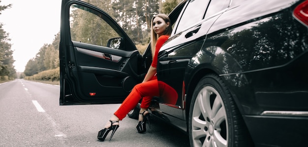 Красивая молодая девушка в красном комбинезоне сидит за рулем черной машины на пустой дороге в лесу