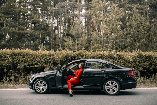 A beautiful young girl in a red overalls sits behind the wheel of a black car on an empty road in the forest