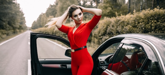 A beautiful young girl in a red overalls stands by a black car on an empty road in the forest