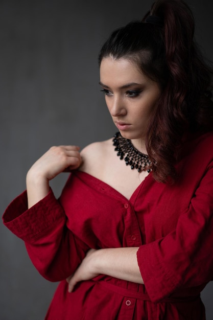 A beautiful young girl in a red shirt with a necklace with a handmade ornament on her neck is posing in a photo studio