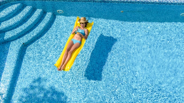 Beautiful young girl relaxing in swimming pool, swims on inflatable mattress and has fun in water on family vacation, tropical holiday resort, aerial drone view from above