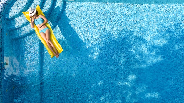 Beautiful young girl relaxing in swimming pool, swims on inflatable mattress and has fun in water on family vacation, tropical holiday resort, aerial drone view from above