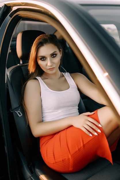 A beautiful young girl sits in a car in the evening in the sunset summer sun in an empty parking lot