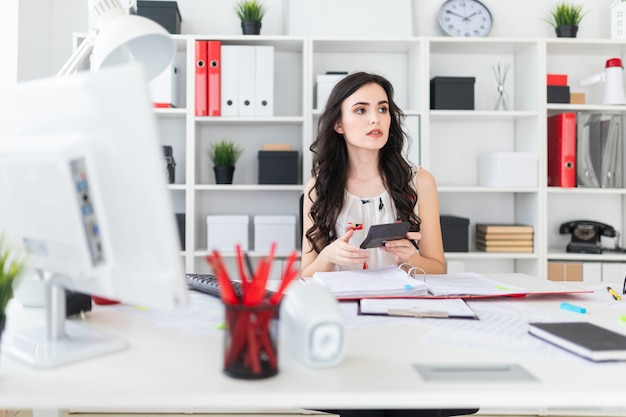 Beautiful young girl sits at the office desk and holds a pen and a calculator in her hands.