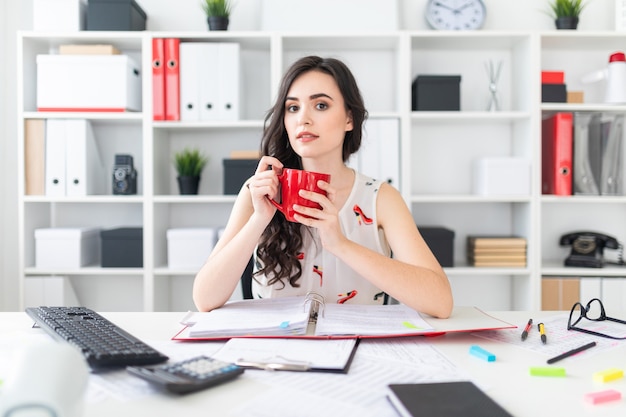 美しい少女はオフィスの机に座って、手に赤いマグカップを保持しています。