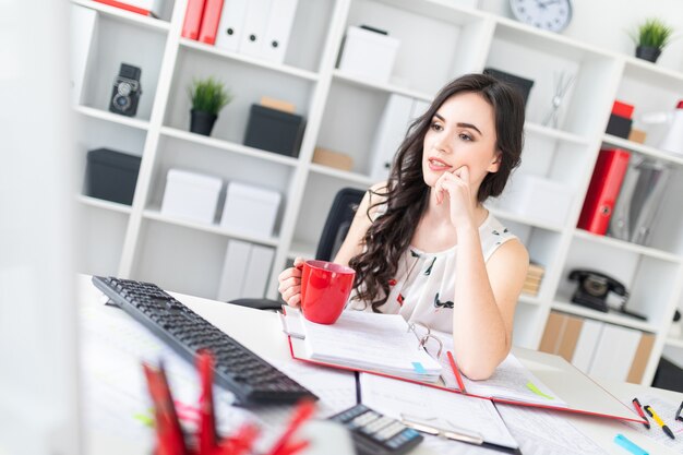 Beautiful young girl sits at office desk, looks at computer screen and holds a red mug in hands.