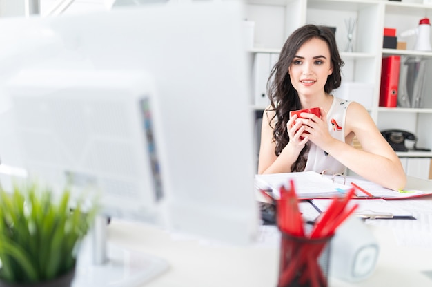 Красивая молодая девушка сидит на офисном столе, смотрит на экран компьютера и держит в руках красную кружку.