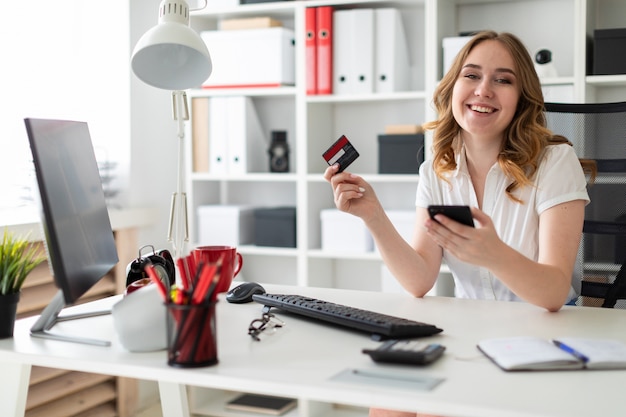 Beautiful young girl sits in the office, holds a bank card and phone in her hand.