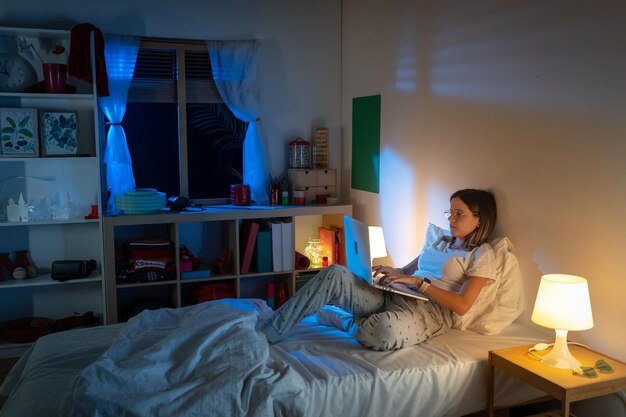 Красивая молодая девушка сидит в постели по ночам и работает с ноутбуком посреди ночи.