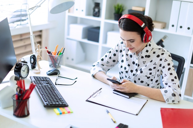 Beautiful young girl sitting in headphones at desk in office and holding a phone.