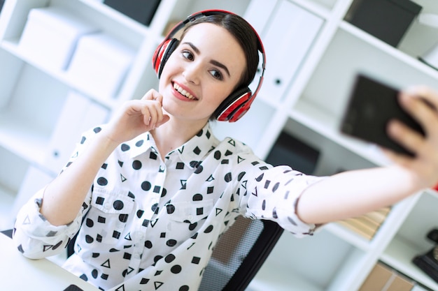 Beautiful young girl sitting in headphones at desk in office and taking pictures of herself on the phone. 