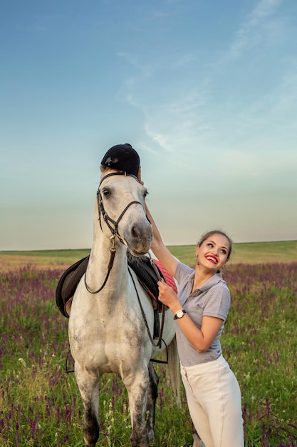 美しい若い女の子は彼女の馬のドレッシングの制服の競争に微笑む：日没の屋外の肖像画。動物の世話、愛と友情の概念。