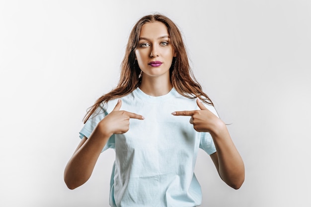 A beautiful young girl smiles and points her fingers at herself on a white isolated gray background. Place for advertising. Positive woman with fashion make-up with purple lips.