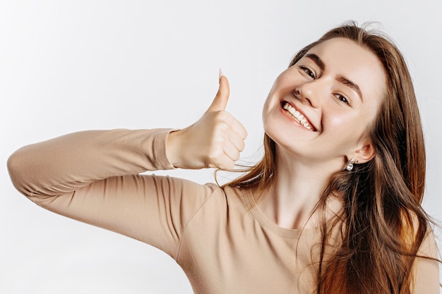 La bella ragazza che sorride e mostra i pollici aumenta il gesto su uno spazio isolato bianco