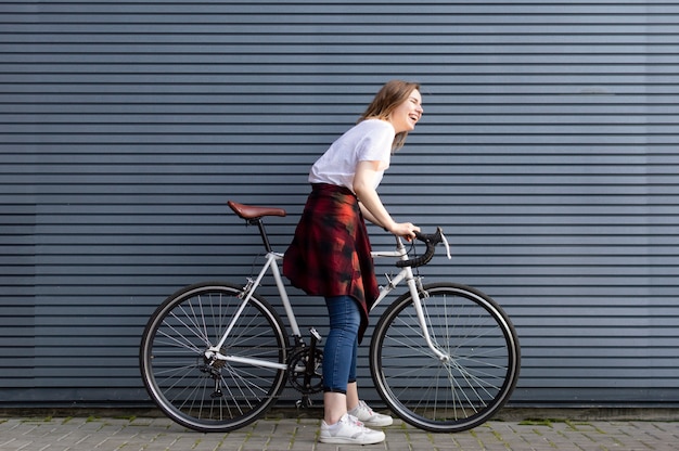 Beautiful young girl standing with a white bicycle on background of gray striped wall