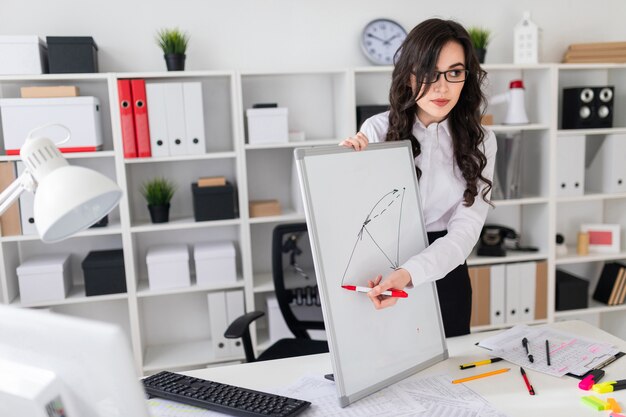 A beautiful young girl stands near an office desk and draws a magnetic marker on the magnetic board.