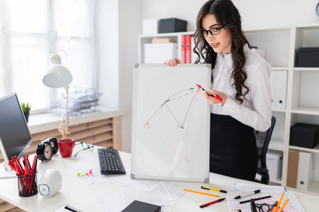 A beautiful young girl stands near an office desk and draws a magnetic marker on the magnetic board.