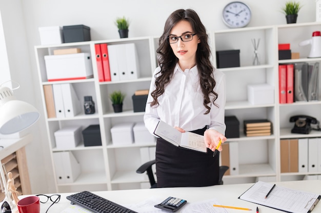 A beautiful young girl stands near an office desk and holds a pen and a notebook in her hands. The girl is negotiating.