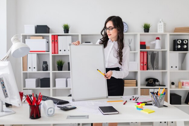 Beautiful young girl stands near an office desk and points with a pen on an empty board