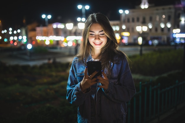 beautiful young girl texting on cell phone outdoor over blurred night street wall, selective focus