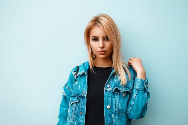 Beautiful young girl in trendy stylish denim clothes near blue wall