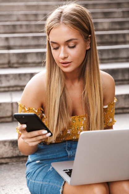 Красивая молодая девушка с помощью портативного компьютера, сидя на лестнице и используя мобильный телефон на открытом воздухе