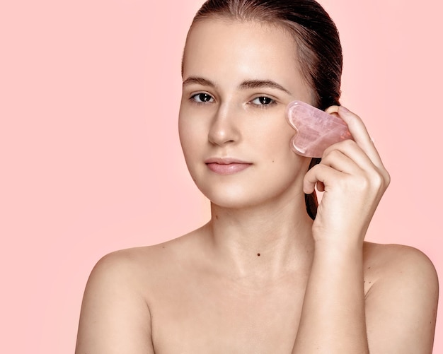 クォーツ・グア・シャ・ファイシャル・スクラバーを使用した美しい若い女の子がスキンケアコンセプトで顔の皮膚をマッサージしています