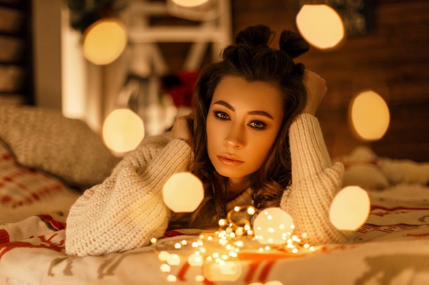 Bella ragazza in un maglione vintage con luci natalizie che riposa sul letto