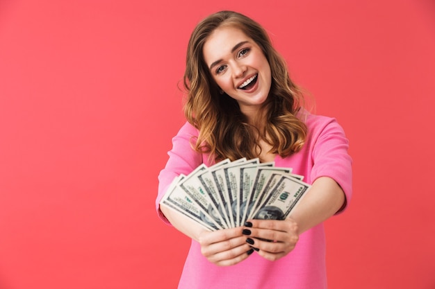 Beautiful young girl wearing casual clothes standing isolated over pink wall, showing money banknotes