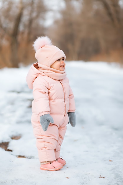 A beautiful young girl wearing a pink jumpsuit running in a snowy winter park 