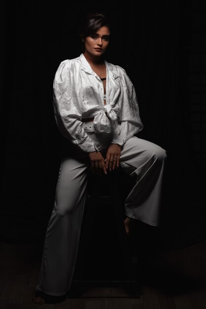 Beautiful young girl in white clothes posing in a dark photo studio. Low key photo. Sensitive brown-haired woman with long hair