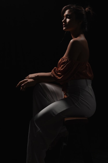 Beautiful young girl in white clothes posing in a dark photo studio. Low key photo. Sensitive brown-haired woman with long hair