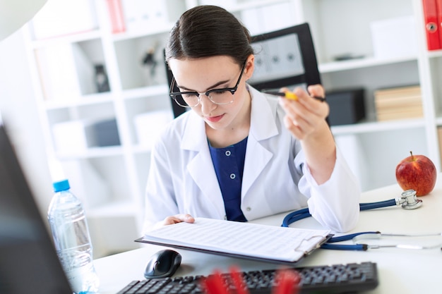 A beautiful young girl in a white robe is sitting at a computer desk with documents and a pen in her hands.