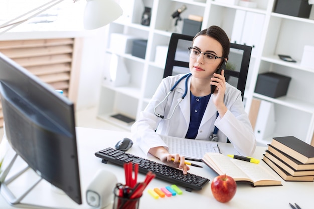 A beautiful young girl in a white robe is sitting at the table, talking on the phone and holding her hand on the keyboard.