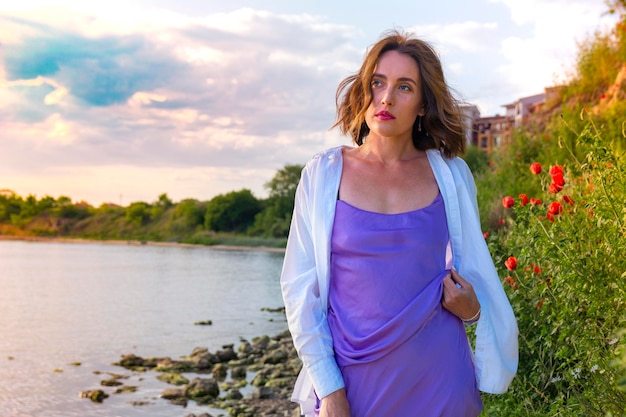 A beautiful young girl in a white shirt and purple dress poses against the backdrop of a pond and blooming poppies