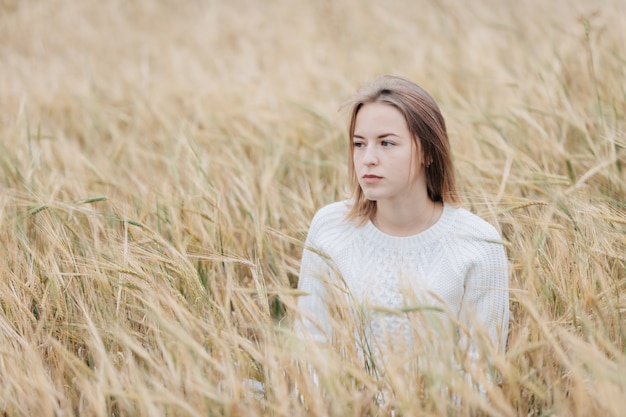 Beautiful young girl in a white sweater sits in a wheat field.