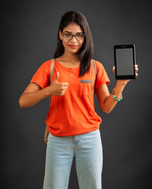 A beautiful young girl with a backpack shows a blank screen of a smartphone or mobile or tablet phone posing on a grey background