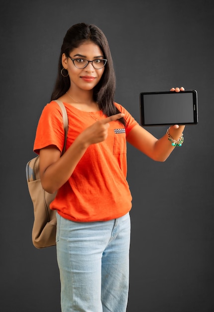 Una bella ragazza con uno zaino mostra uno schermo vuoto di uno smartphone o un telefono cellulare o tablet in posa su uno sfondo grigio