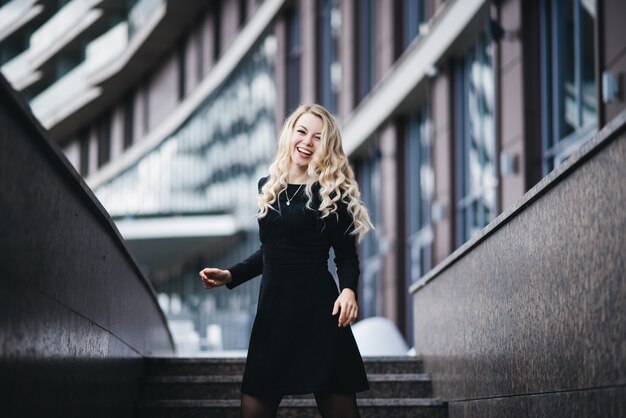 Beautiful young girl with blond wavy hair in black dress having fun on a  modern building