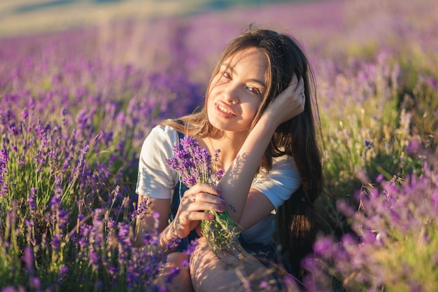 Beautiful young girl with a bouquet of flowers sits in a lavender field in the sunlight