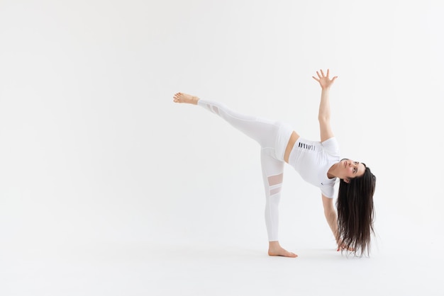a beautiful young girl with dark hair stands in the pose of Ardha Chandrasana on a white background