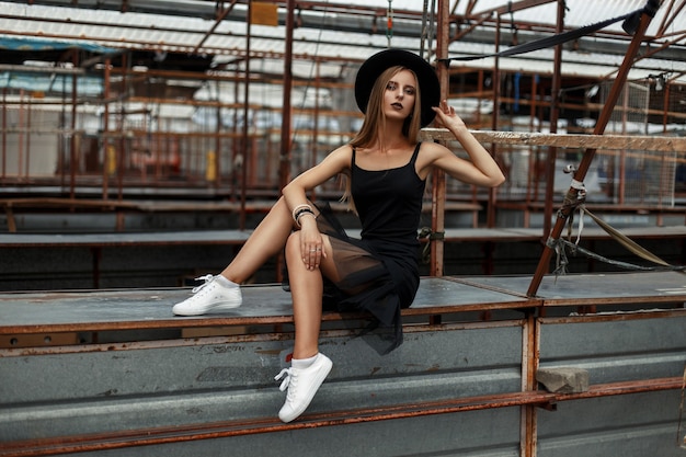 Beautiful young girl with fashionable black clothes with a hat and white shoes sitting on a metal surface